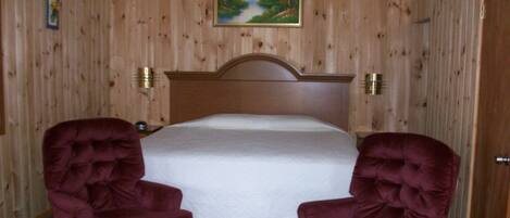 Un dormitorio con pared de paneles de madera, una cama con colchón blanco y dos sillones tapizados.