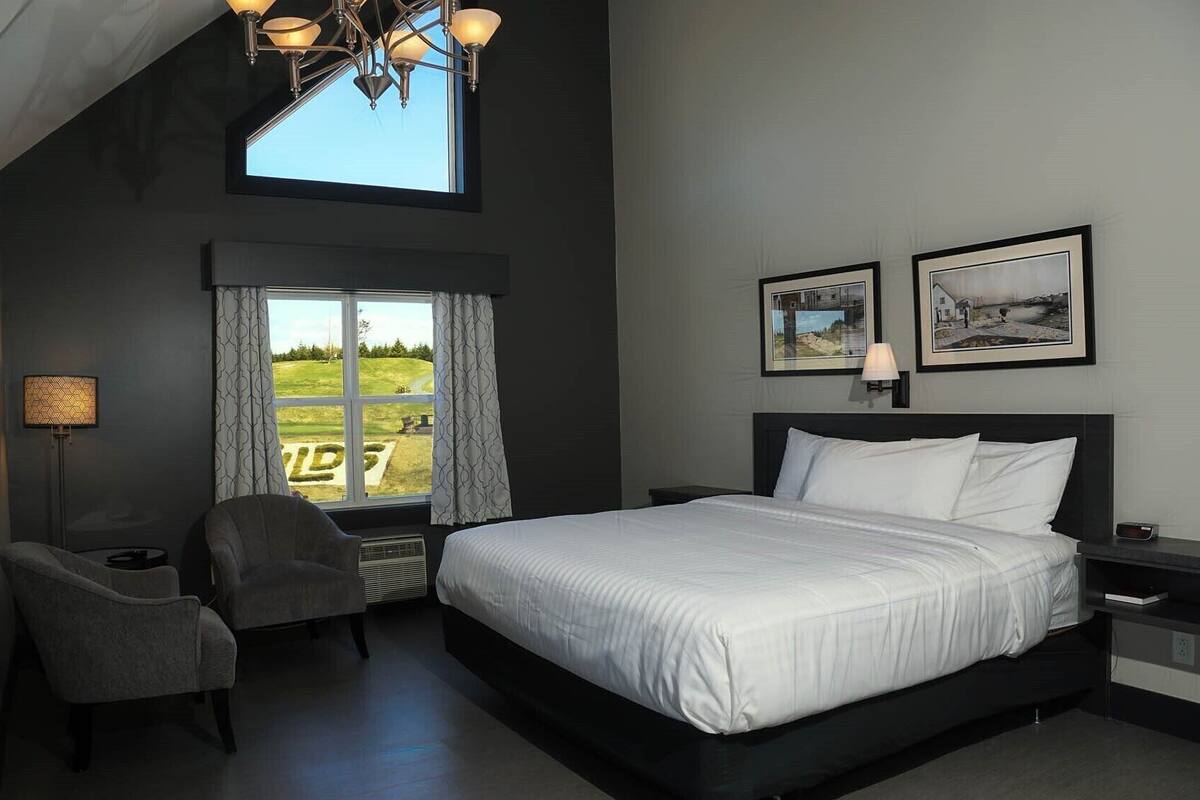 A modern bedroom with a large bed, a desk, and a chair. There is a window with curtains and a view of a green landscape.