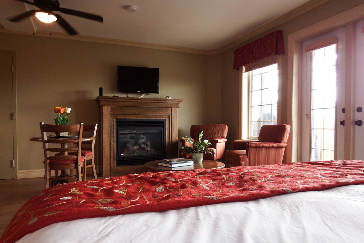 A bedroom with a fireplace, a television, a bed with a red blanket, and a dining area with chairs and a table.