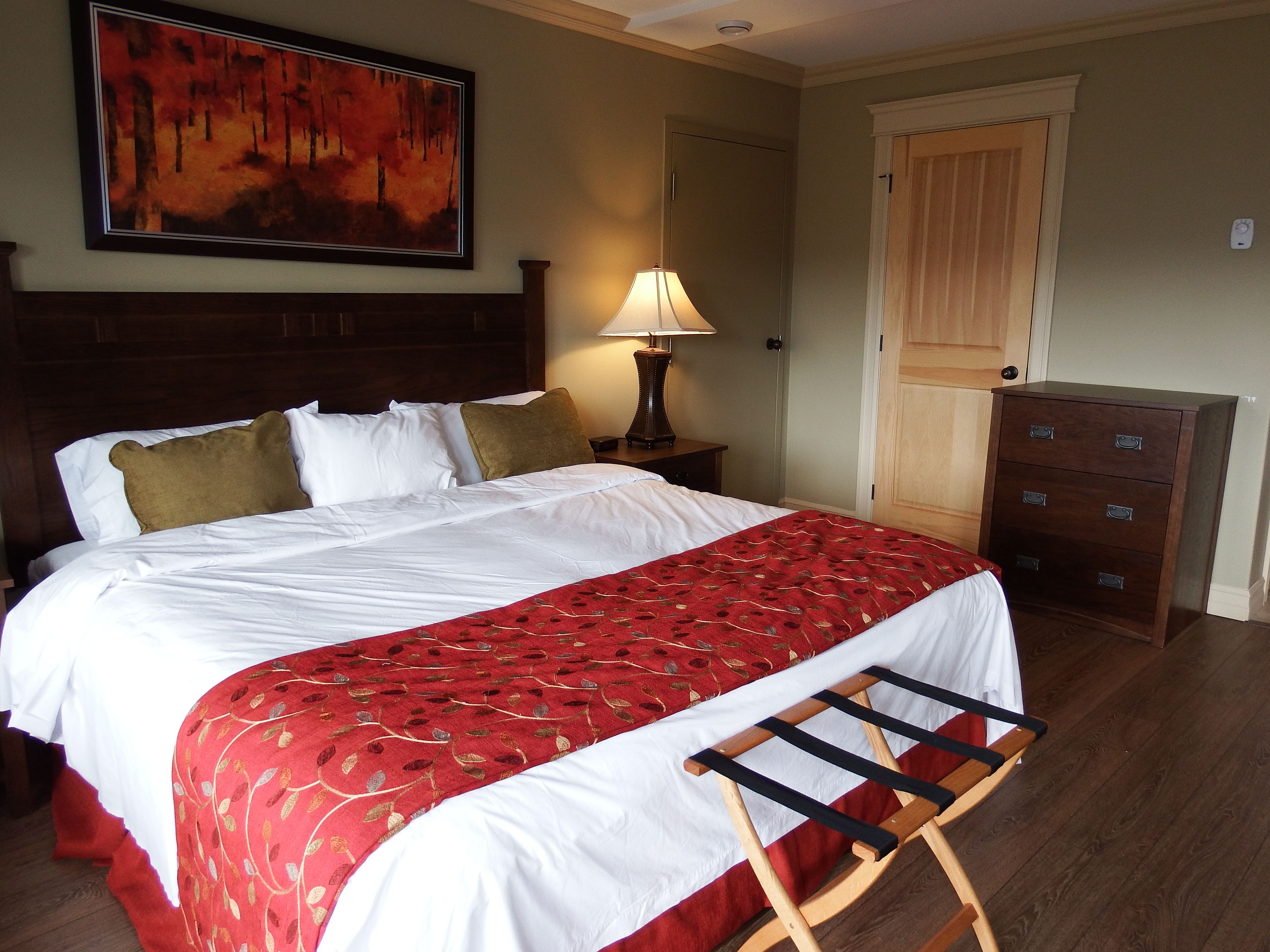 Un lit bien fait, avec une literie blanche et rouge, une tête de lit en bois, une table de chevet avec une lampe, une commode en bois et une porte avec un interrupteur sur le mur.