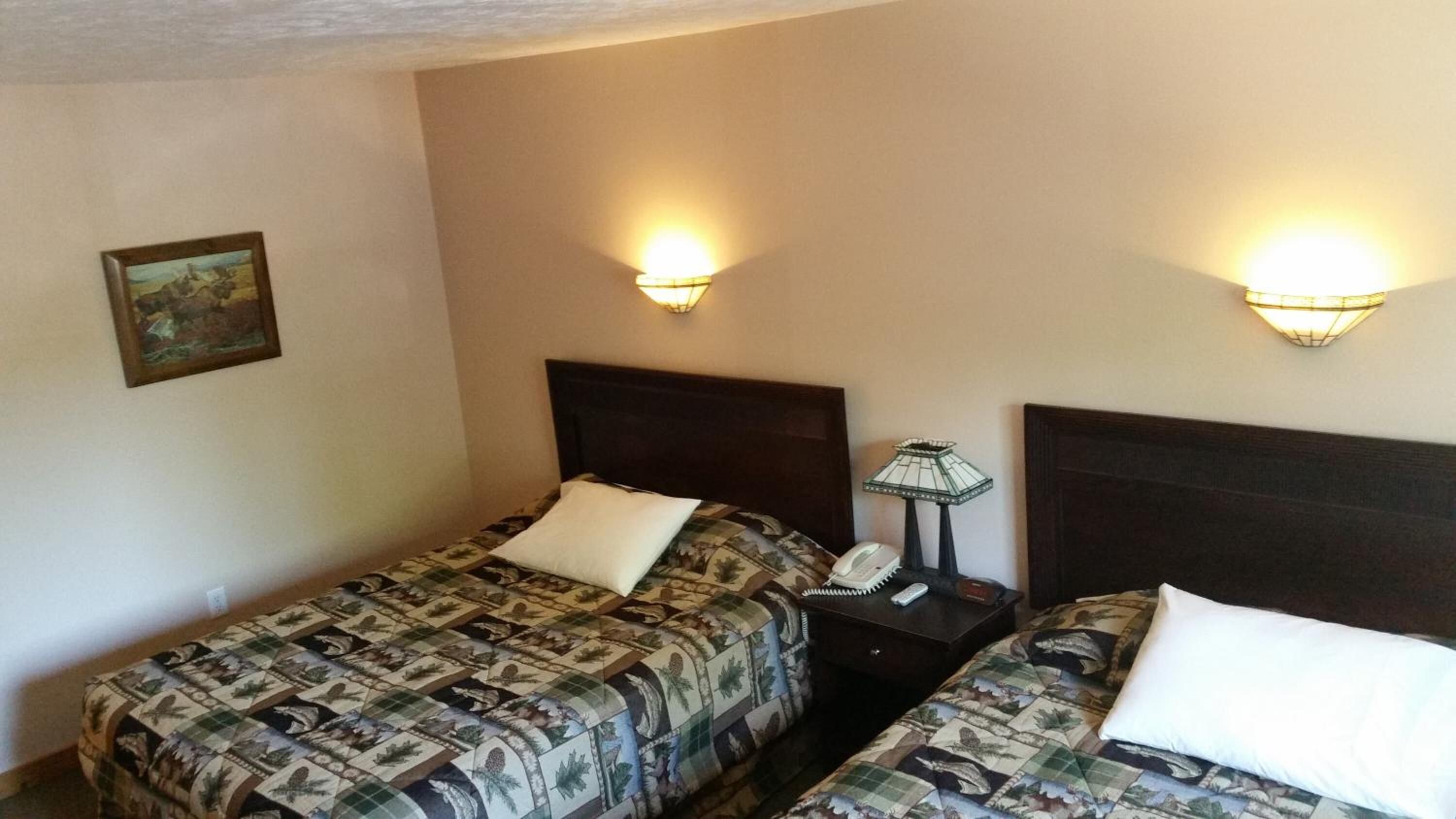 A hotel room with two beds, each with a patterned bedspread and white pillows. There are wall-mounted lamps on either side of the beds.