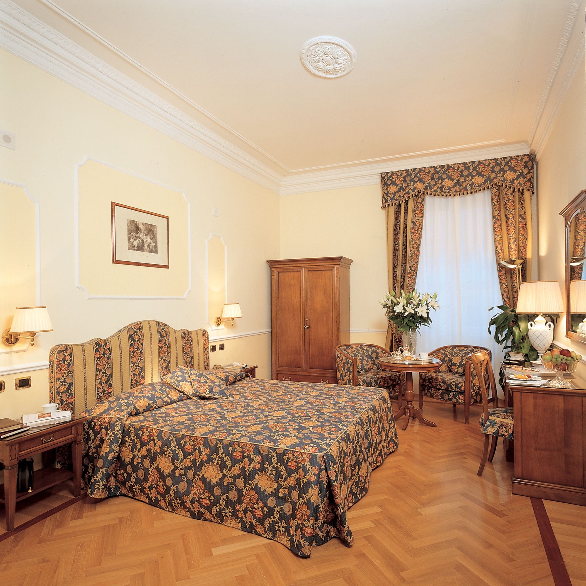 Une chambre d’hôtel avec un grand lit, une armoire en bois, un bureau avec une lampe, une chaise et une petite table sur laquelle est posé un vase de fleurs.