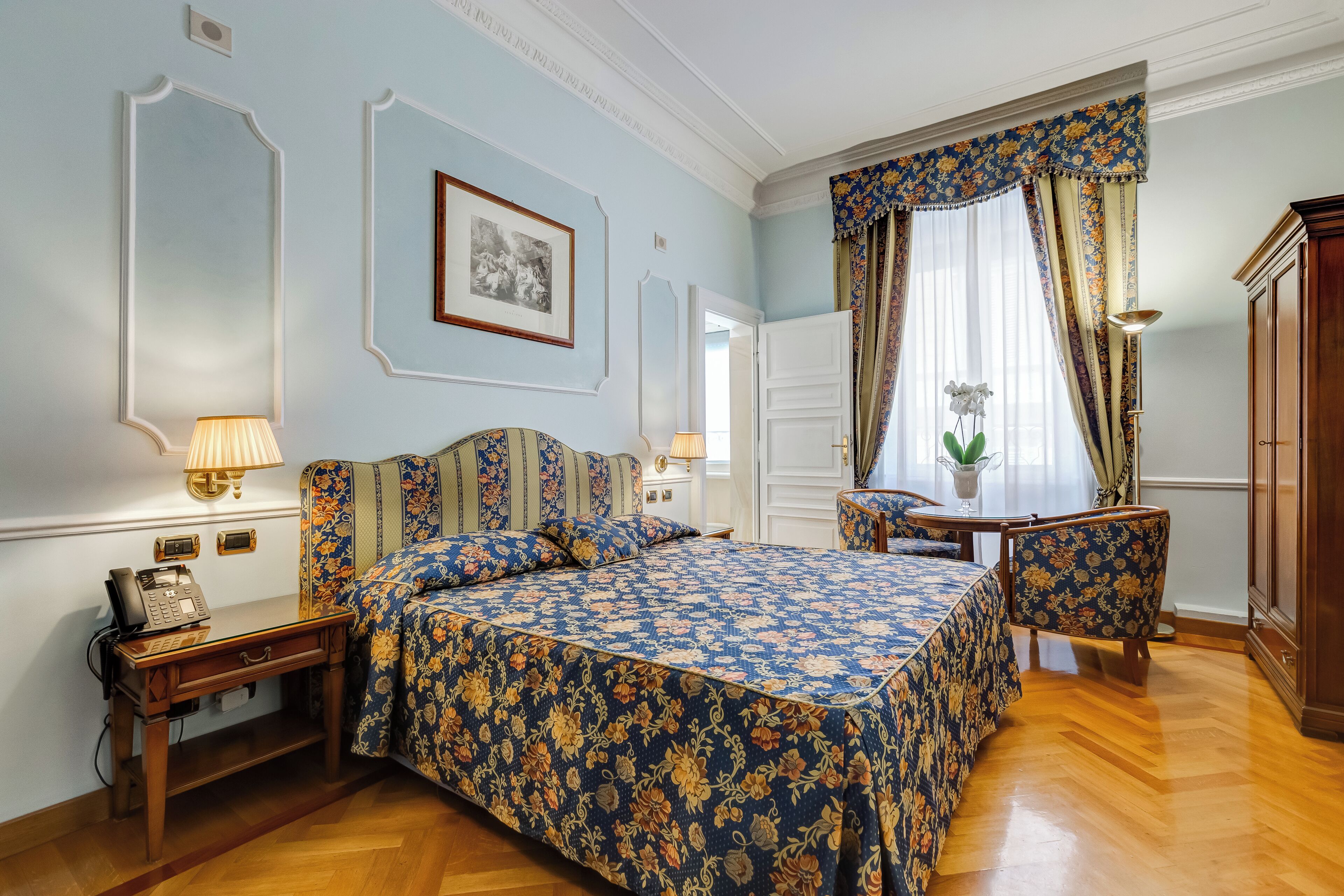 Un dormitorio con colcha estampada, una mesita de noche con lámpara, una silla y un armario de madera.
