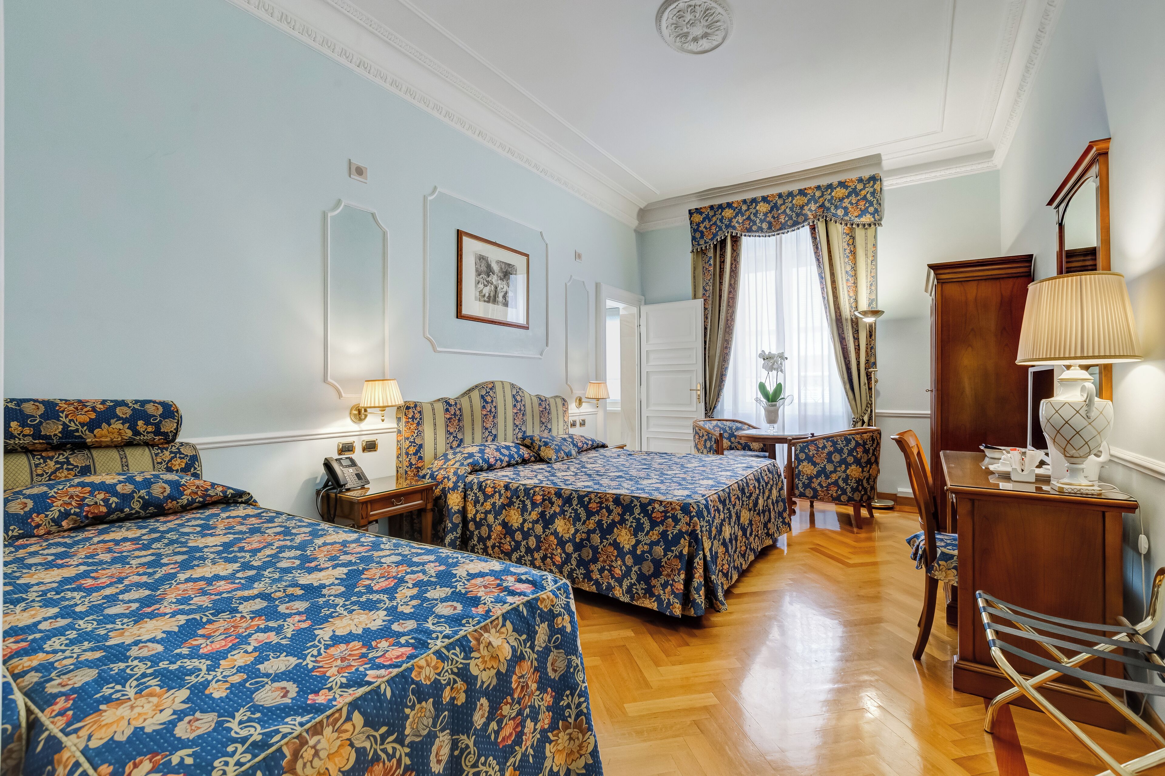 A hotel room with two beds, a desk, a chair, and a lamp.