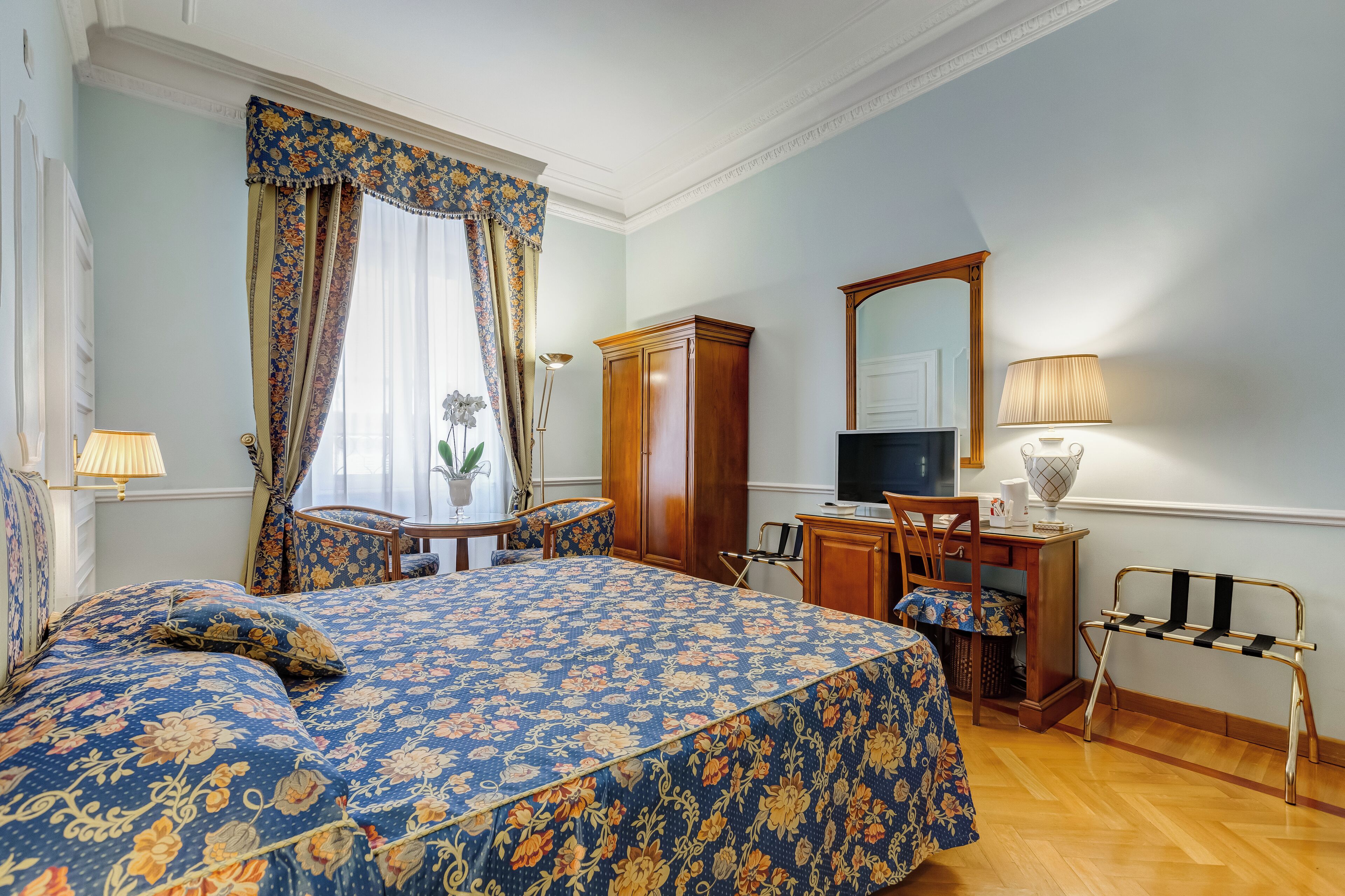 A bedroom with a floral bedspread, a television, a desk with a lamp, and a mirror on the wall.