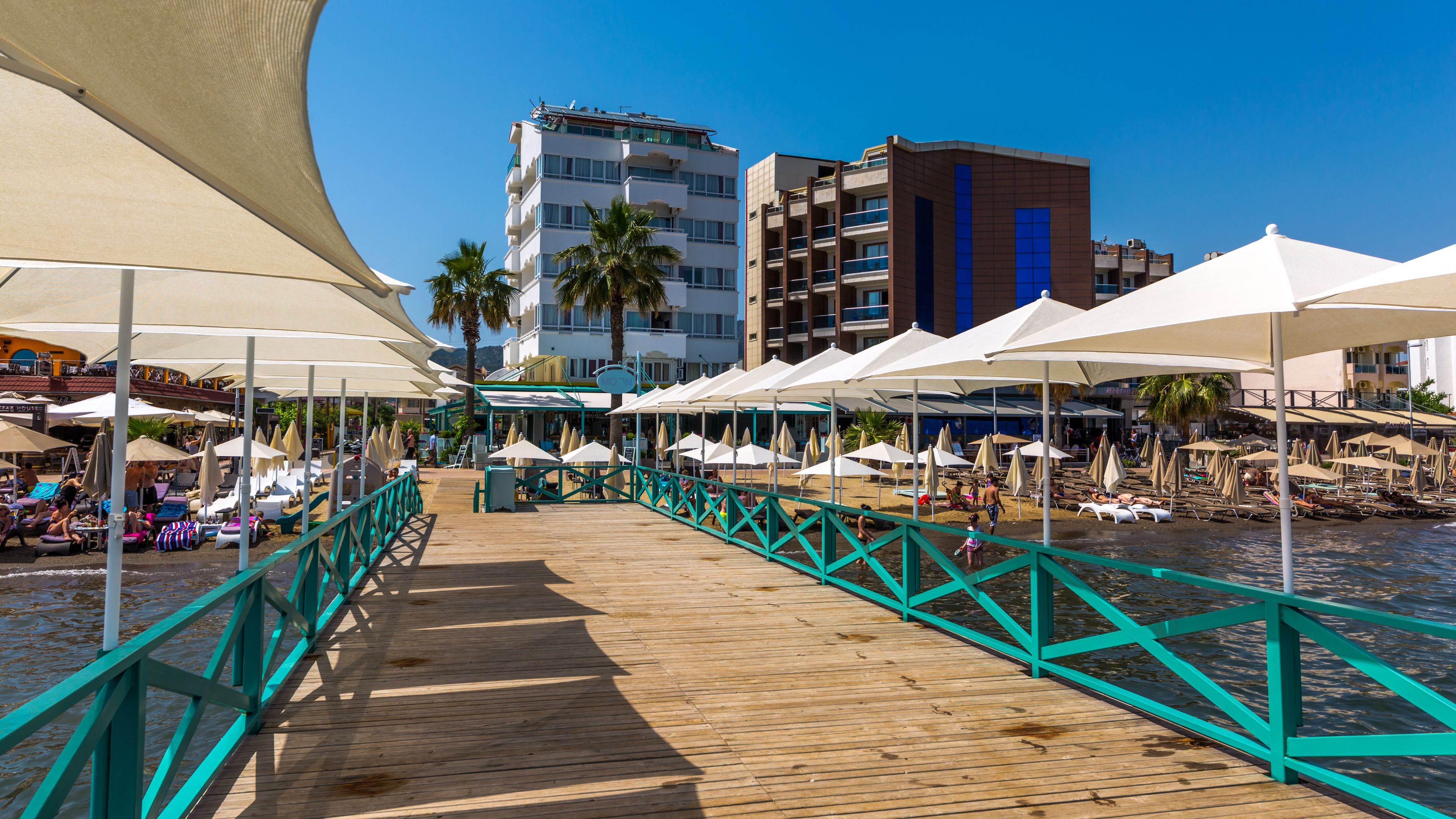 A wooden boardwalk with turquoise railings leading to beach umbrellas and lounge chairs.