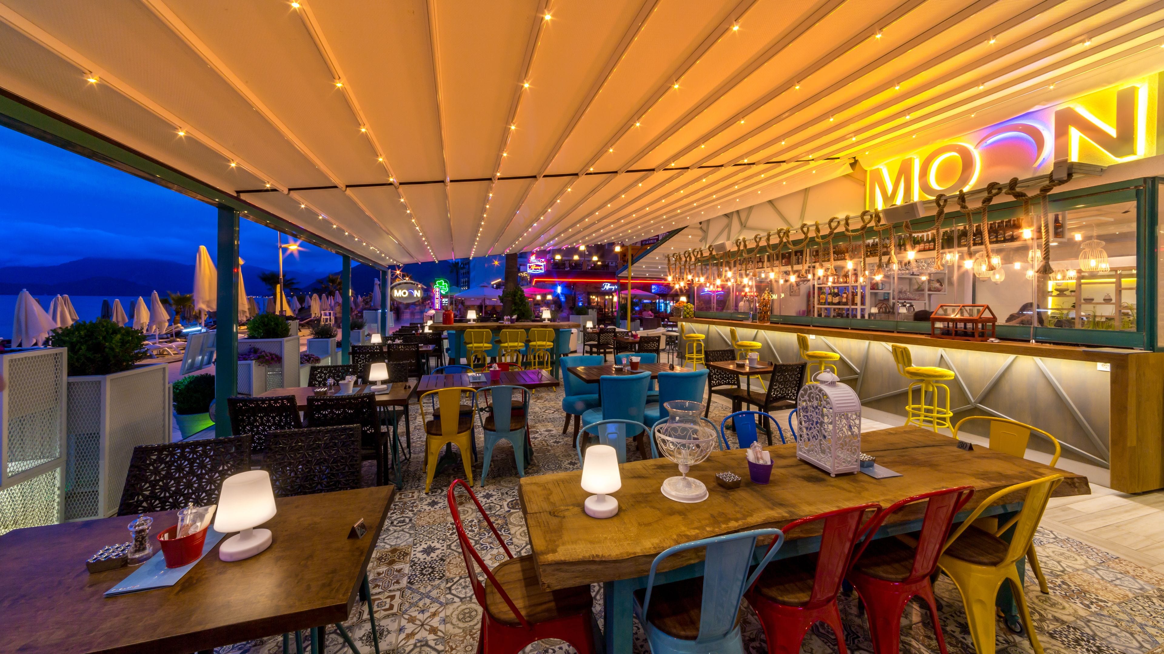 An outdoor dining area with multiple tables and chairs, a bar with a neon sign, and a view of the sea.