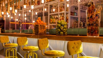 Zona de bar con taburetes amarillos, una exhibición de botellas y un cesto de naranjas.