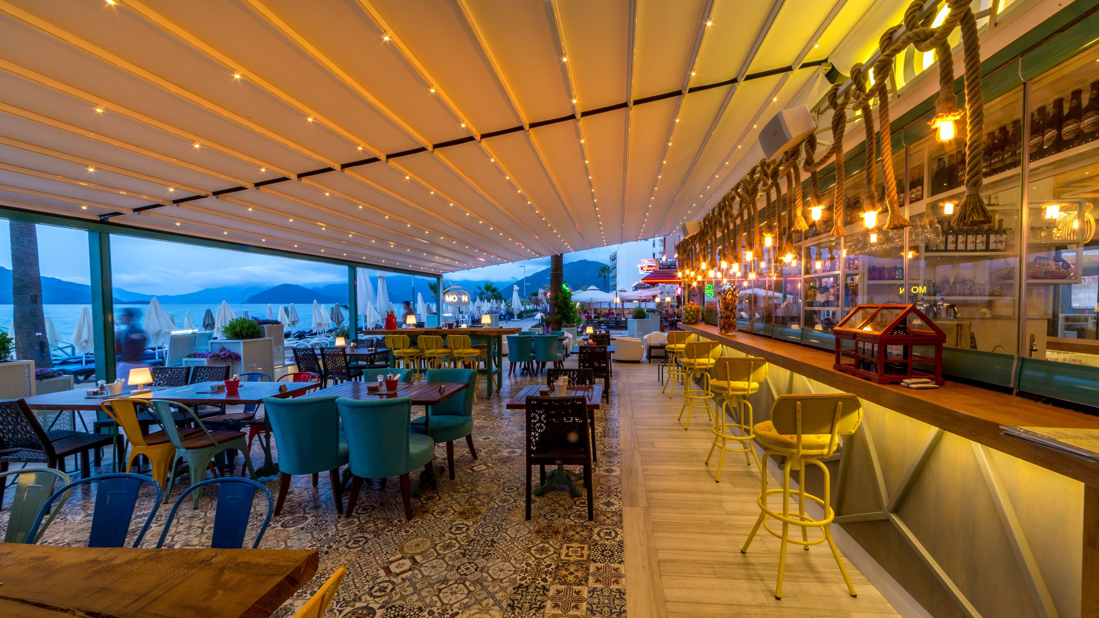 Een restaurant met een terras met uitzicht op het water, met diverse zithoeken en een bar met hangende lampen.
