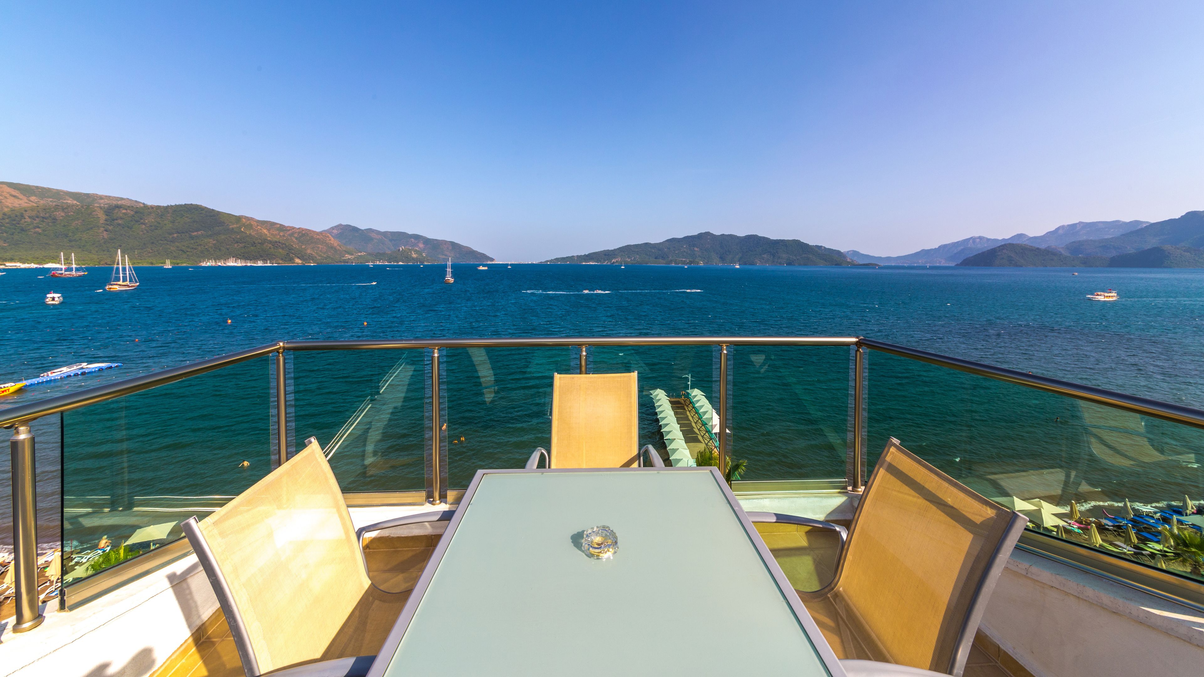 A deck with a table and chairs overlooking a body of water with boats and distant hills.