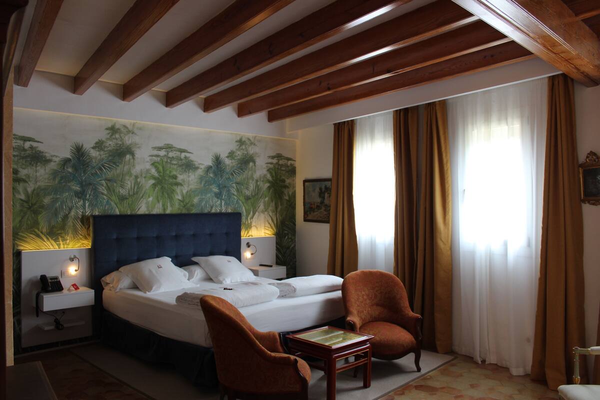 A hotel room with a large bed, two chairs, a nightstand with a lamp, and a painting of palm trees on the wall.