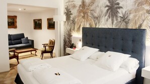 Habitación de hotel con una cama grande, un sofá, una mesita y una silla. Hay un cuadro en la pared y una lámpara en la mesita de noche.
