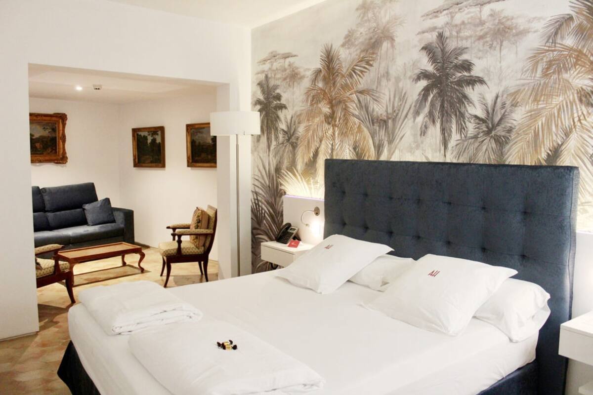 A hotel room with a large bed, a sofa, a small table, and a chair. There is a painting on the wall and a lamp on the side table.