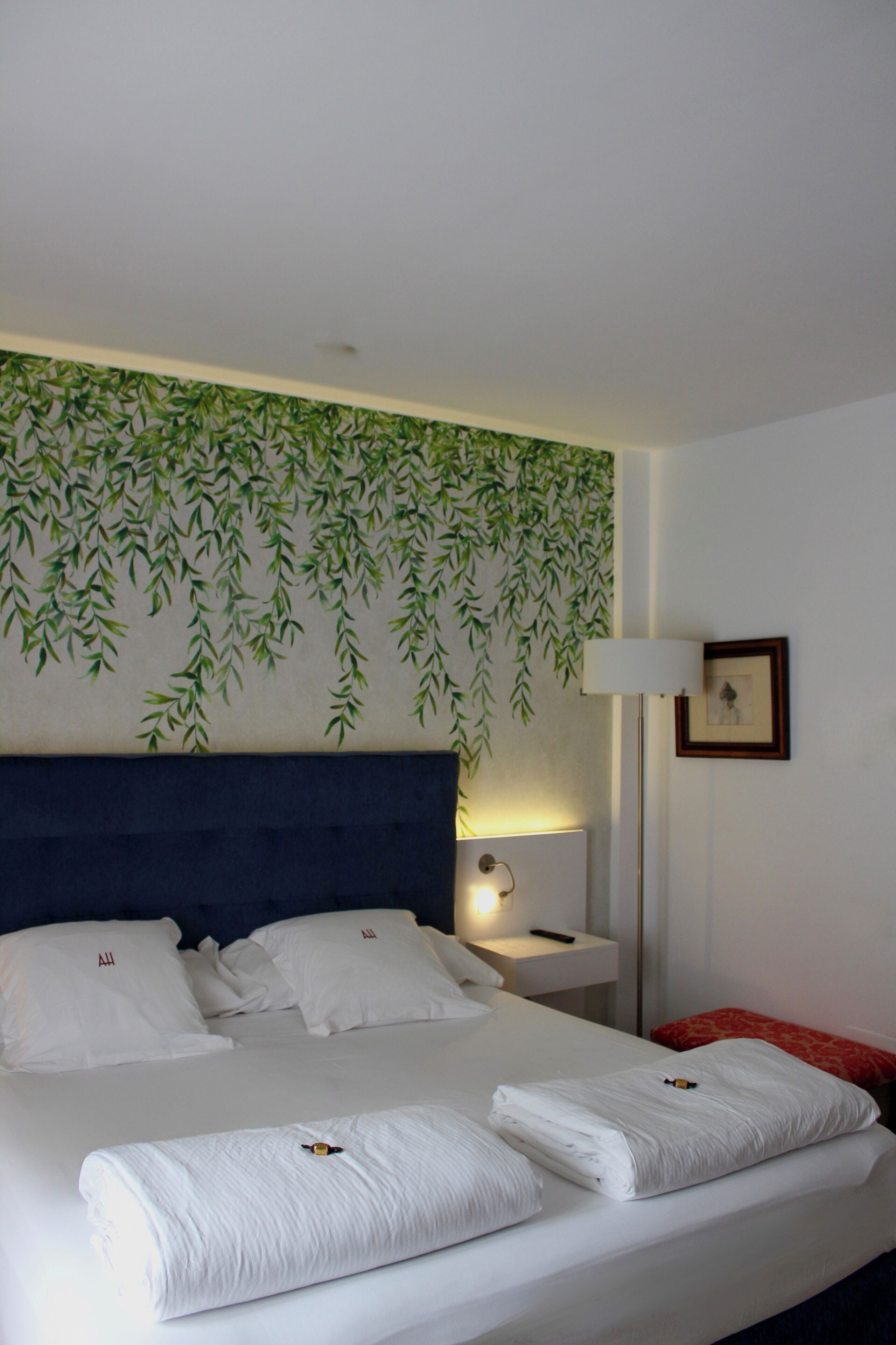A neatly made bed with white linens, a blue headboard, and a wall with a green leafy pattern.