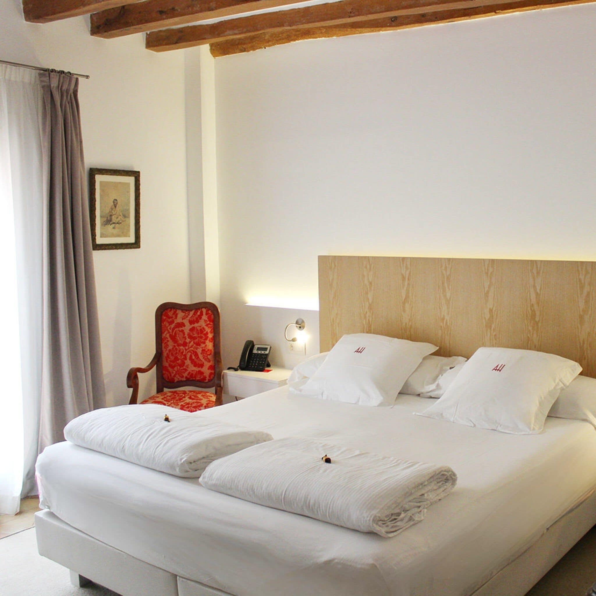 Habitación de hotel con una cama grande, cabecera de madera, una butaca tapizada de rojo, mesita de noche con lámpara y un cuadro enmarcado en la pared.