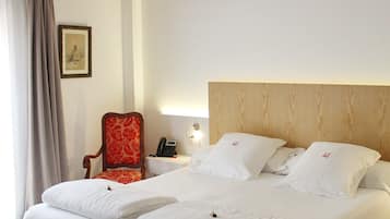 Habitación de hotel con una cama grande, cabecera de madera, una butaca tapizada de rojo, mesita de noche con lámpara y un cuadro enmarcado en la pared.