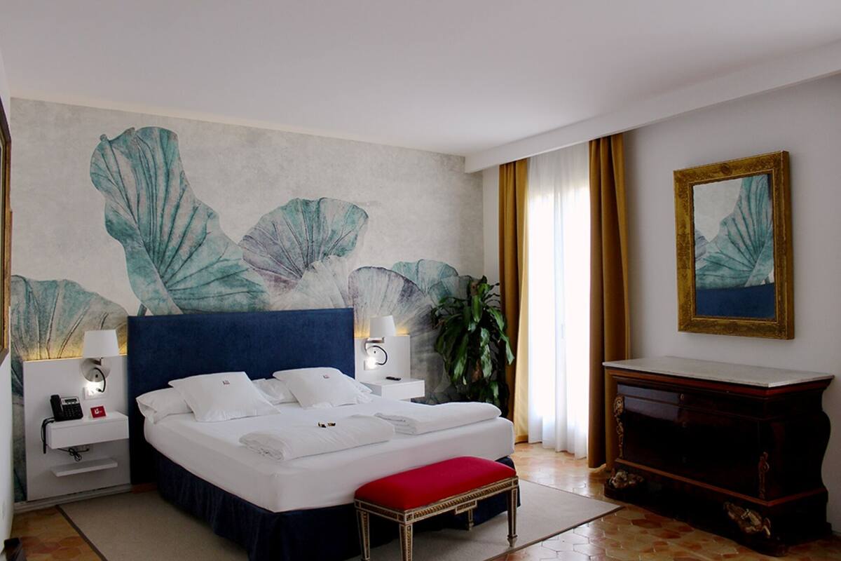 A hotel room with a large bed, a bedside table with a lamp, a small table with a vase, and a large mirror on the wall.