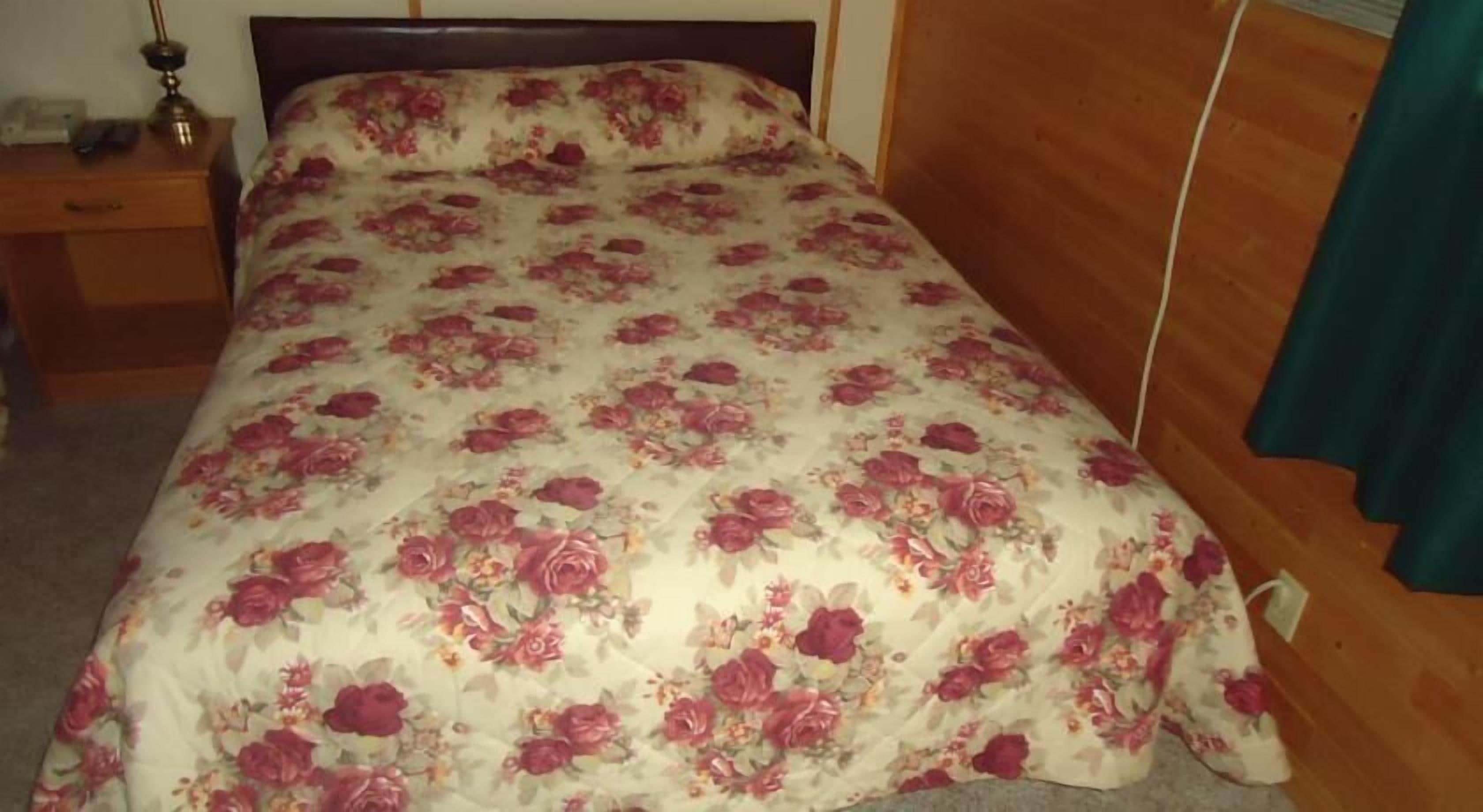 A bed with a floral bedspread, a wooden headboard, a nightstand with a lamp, and a wooden floor.