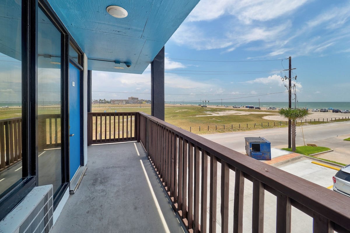 A balcony with a view of a beach, a parking area, and a building in the distance.