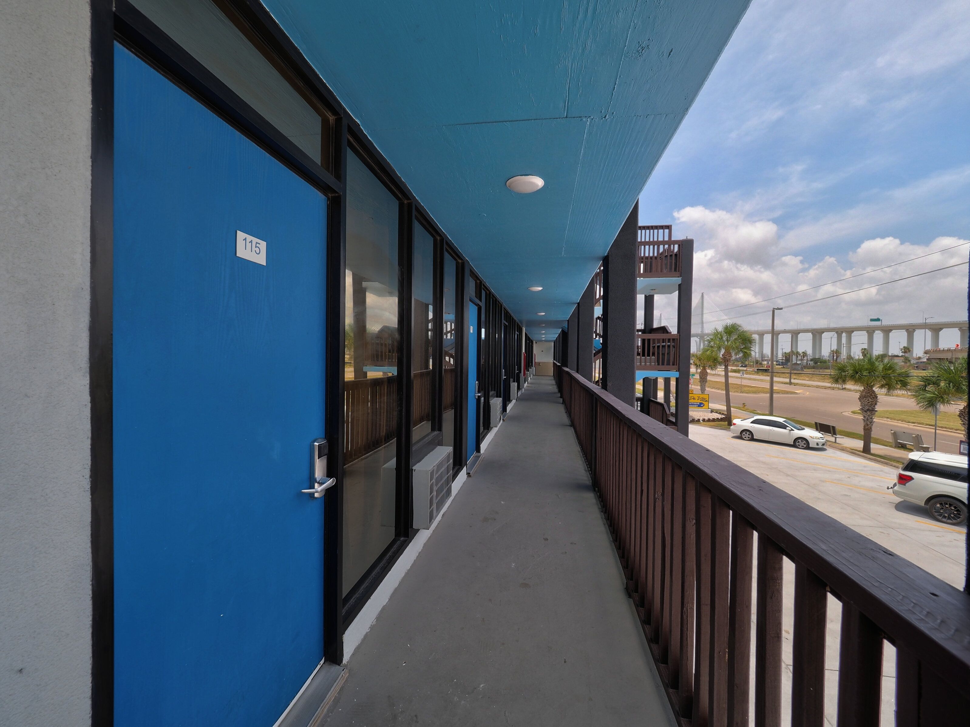 A long corridor with blue doors, a wooden railing, and a view of a bridge and cars in the distance.