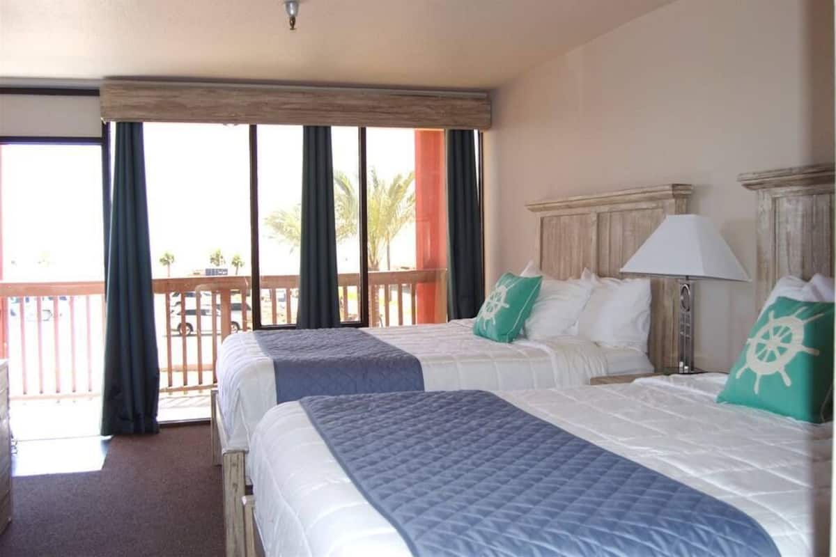 A hotel room with two beds, a balcony with curtains, and a view of palm trees.