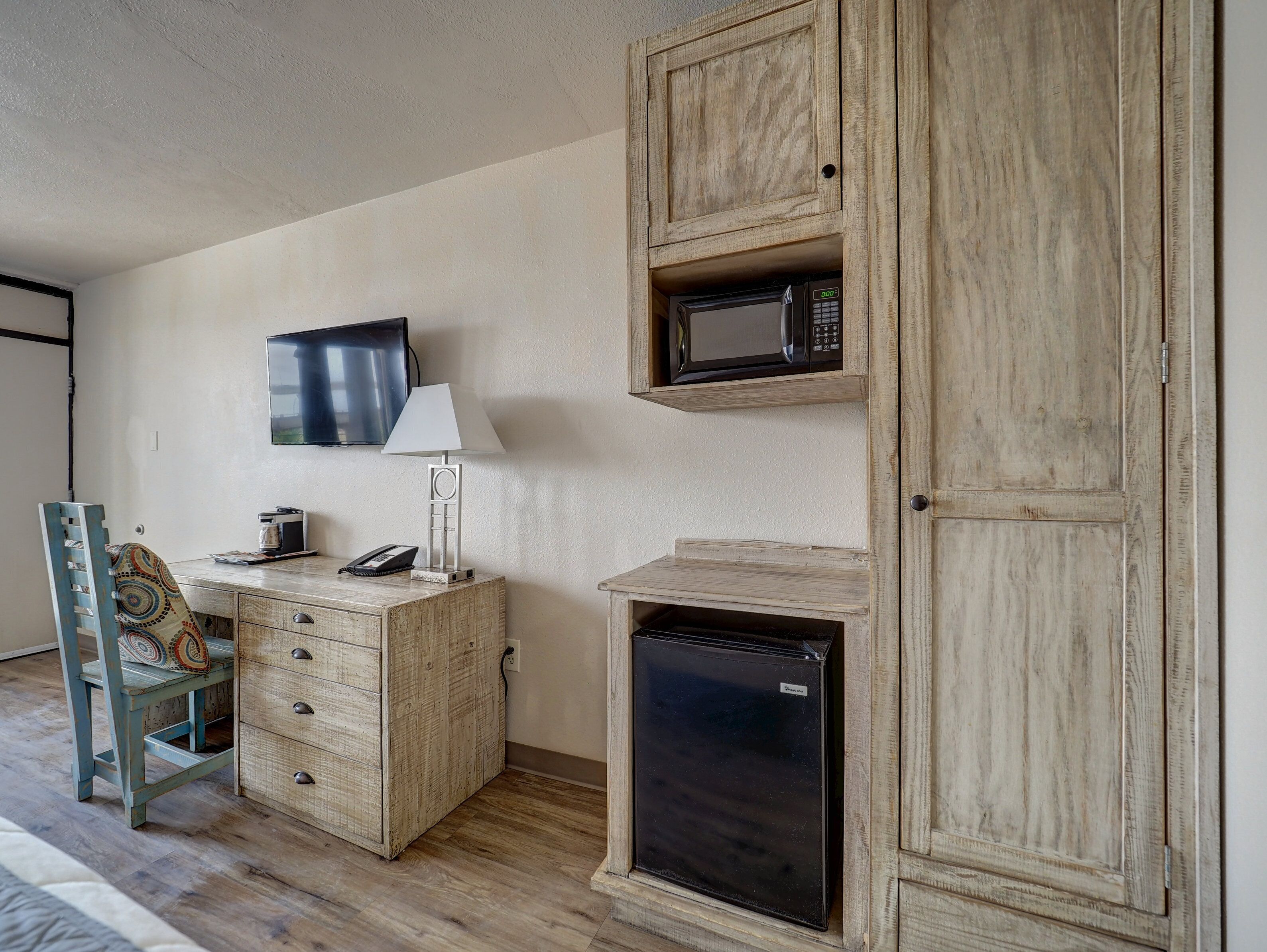 A compact room with a wooden cabinet, a microwave, a small refrigerator, a desk with a lamp, and a television mounted on the wall.