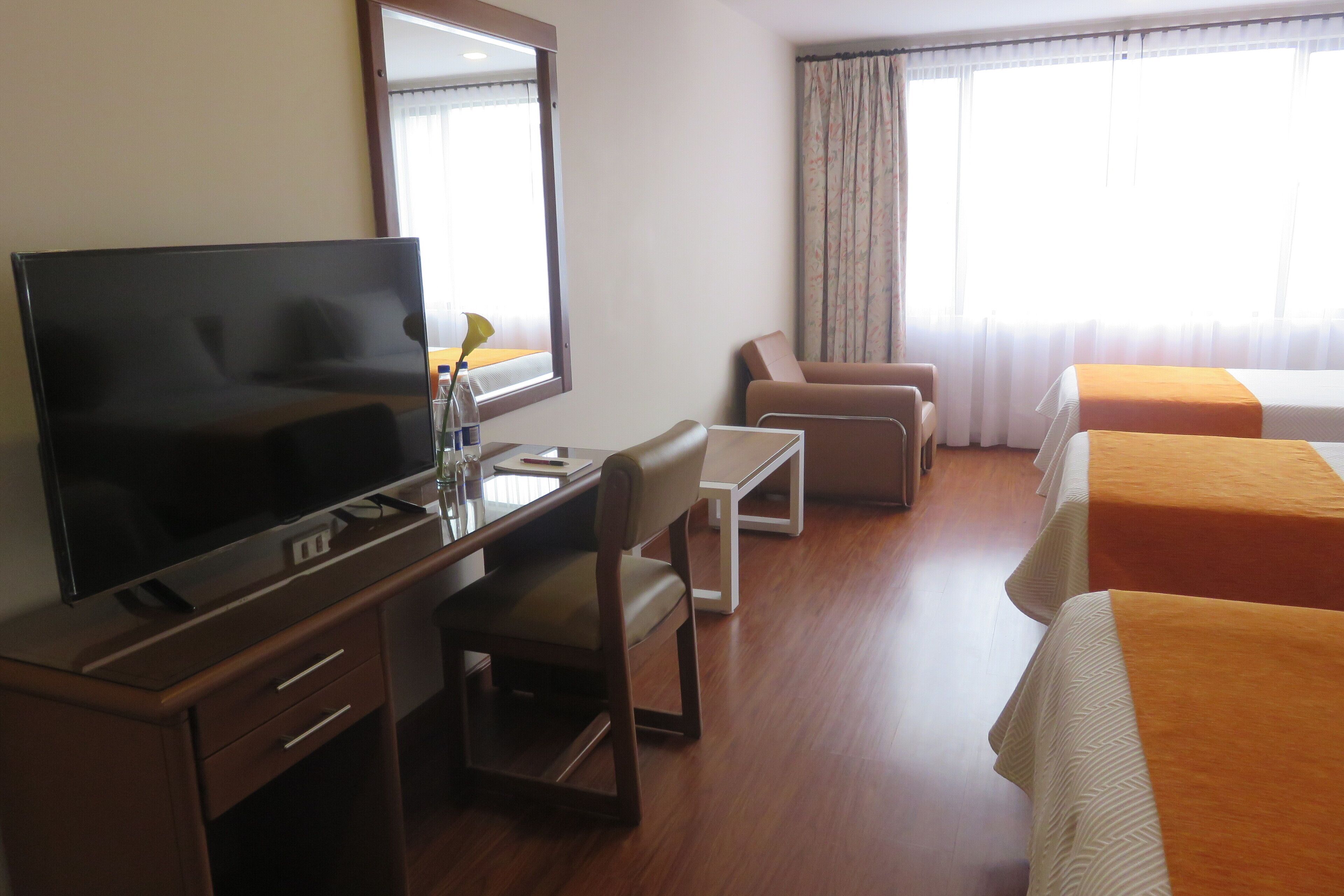Ein Hotelzimmer mit Flachbildfernseher, Schreibtisch, Sessel, einer Couch und zwei Betten mit orangefarbenen Bettdecken.