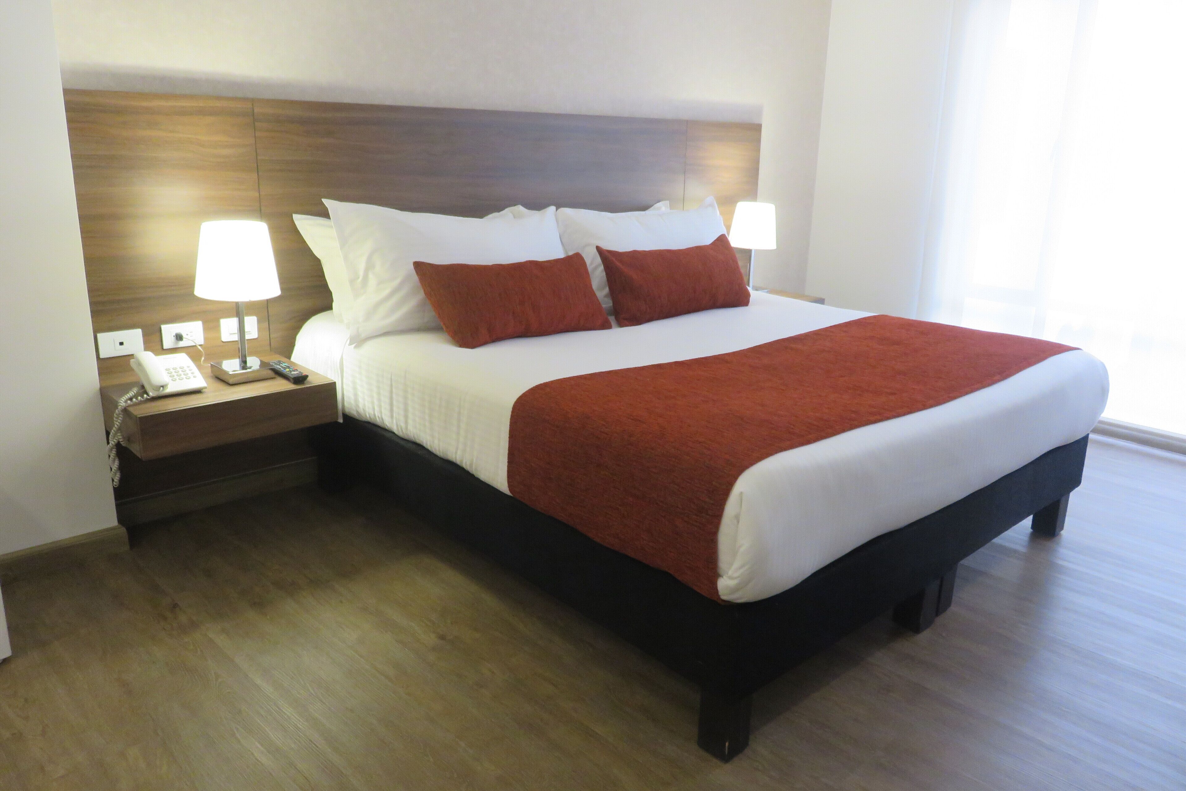 A neatly made bed with a wooden headboard, white linens, and two red pillows. There are bedside lamps on a wooden nightstand, and a telephone.
