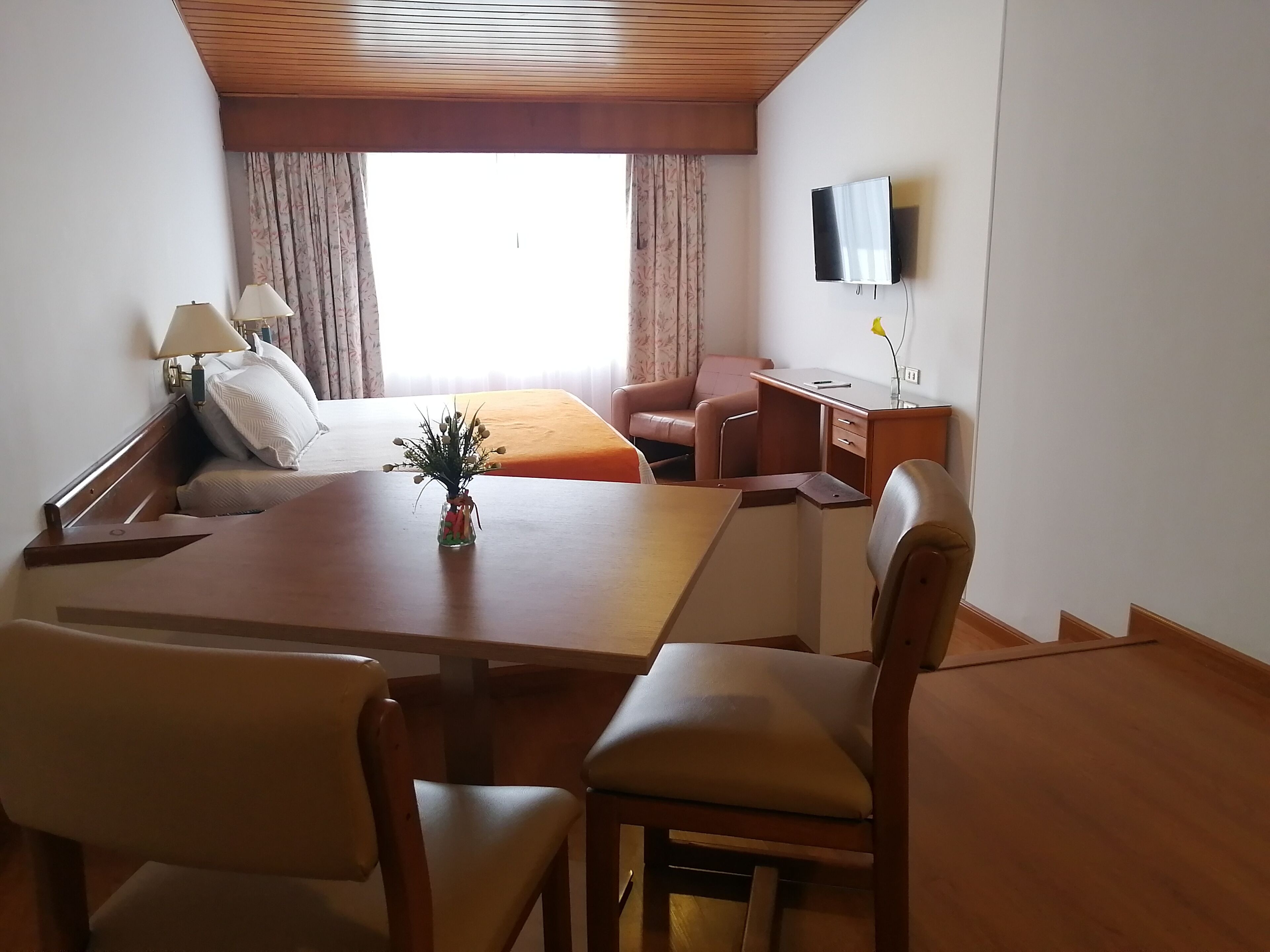 Habitación de hotel con cama, escritorio, silla, televisión y ventana con cortinas.