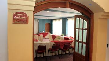 Un umbral abierto que da a un comedor con mesas dispuestas para una comida, un cartel que indica que la sala es parte del Auditorio Iguaque, y vista de la sala contigua con una ventana.