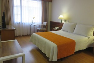 Quarto de hotel com cama, escrivaninha, cadeira, luminária e telefone.
