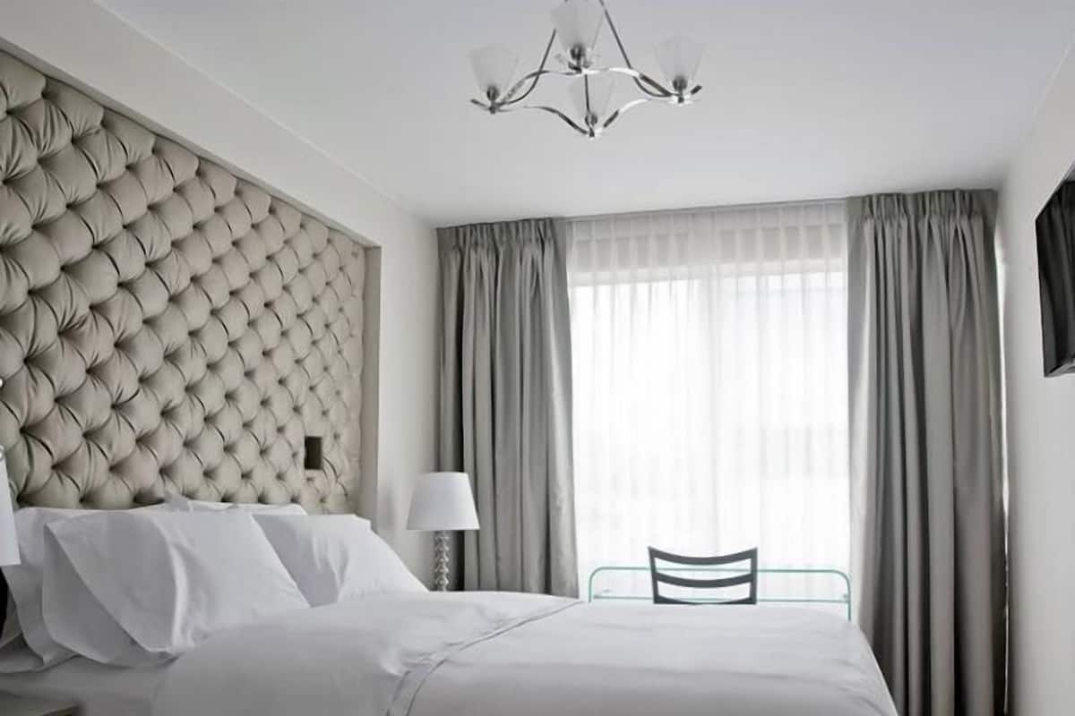 A modern bedroom with a tufted headboard, a bed with white linens, a bedside table with a lamp, and a view of the outside through large windows with curtains.