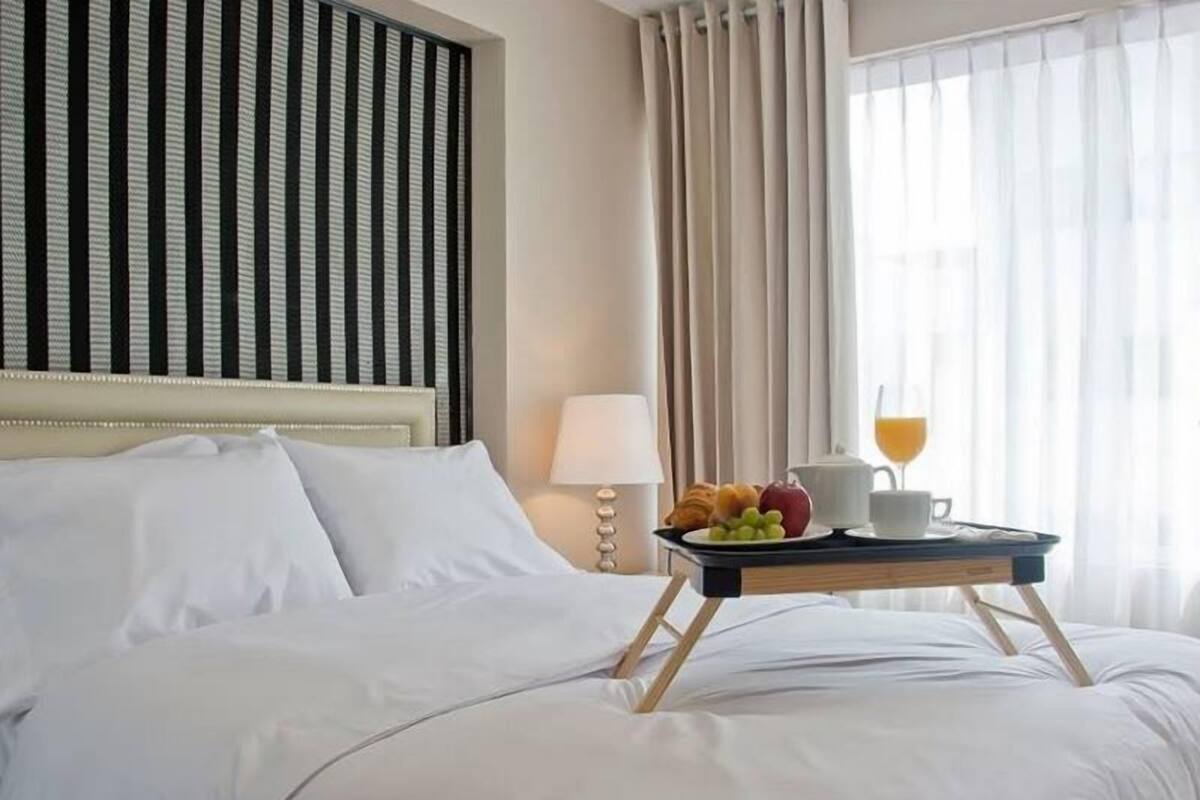 A hotel room with a bed, a bedside table with a lamp, a tray with breakfast items, and a window with curtains.