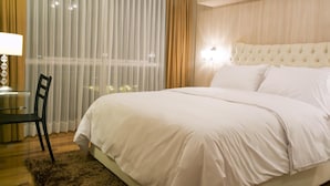 Habitación de hotel con una cama grande, un escritorio con una silla, una lámpara y vistas a la ciudad a través de cortinas translúcidas.