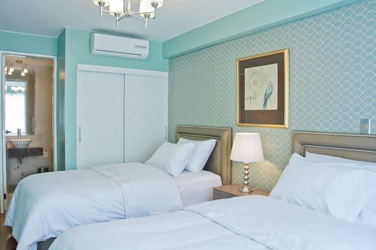 A hotel room with two beds, a bedside table with a lamp, a framed picture on the wall, and a bathroom visible through an open door.