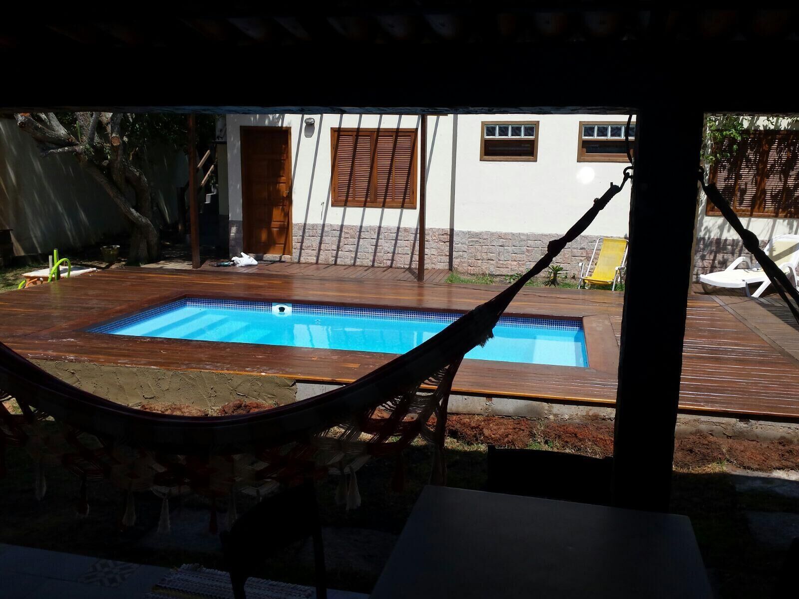 Une piscine rectangulaire entourée d’une terrasse en bois, un hamac et, au fond, un bâtiment doté de fenêtres et d’une porte.