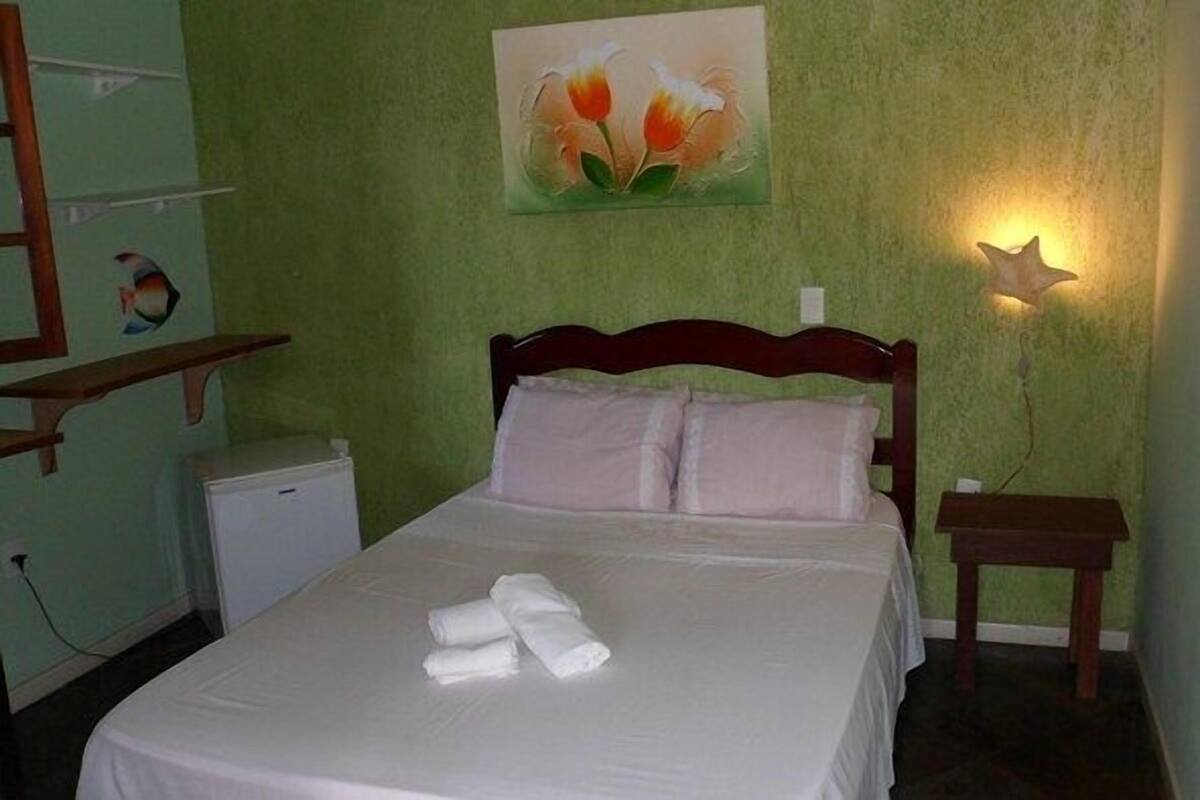 A neatly made bed with pillows and towels, a small bedside table, a nightstand, a wall-mounted lamp, and a painting of flowers on the wall.