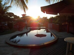 A circular pool with a wooden deck surrounding it, a palm tree, and a building with a red roof in the background. - Pousada Enseada das Ancoras (Búzios)