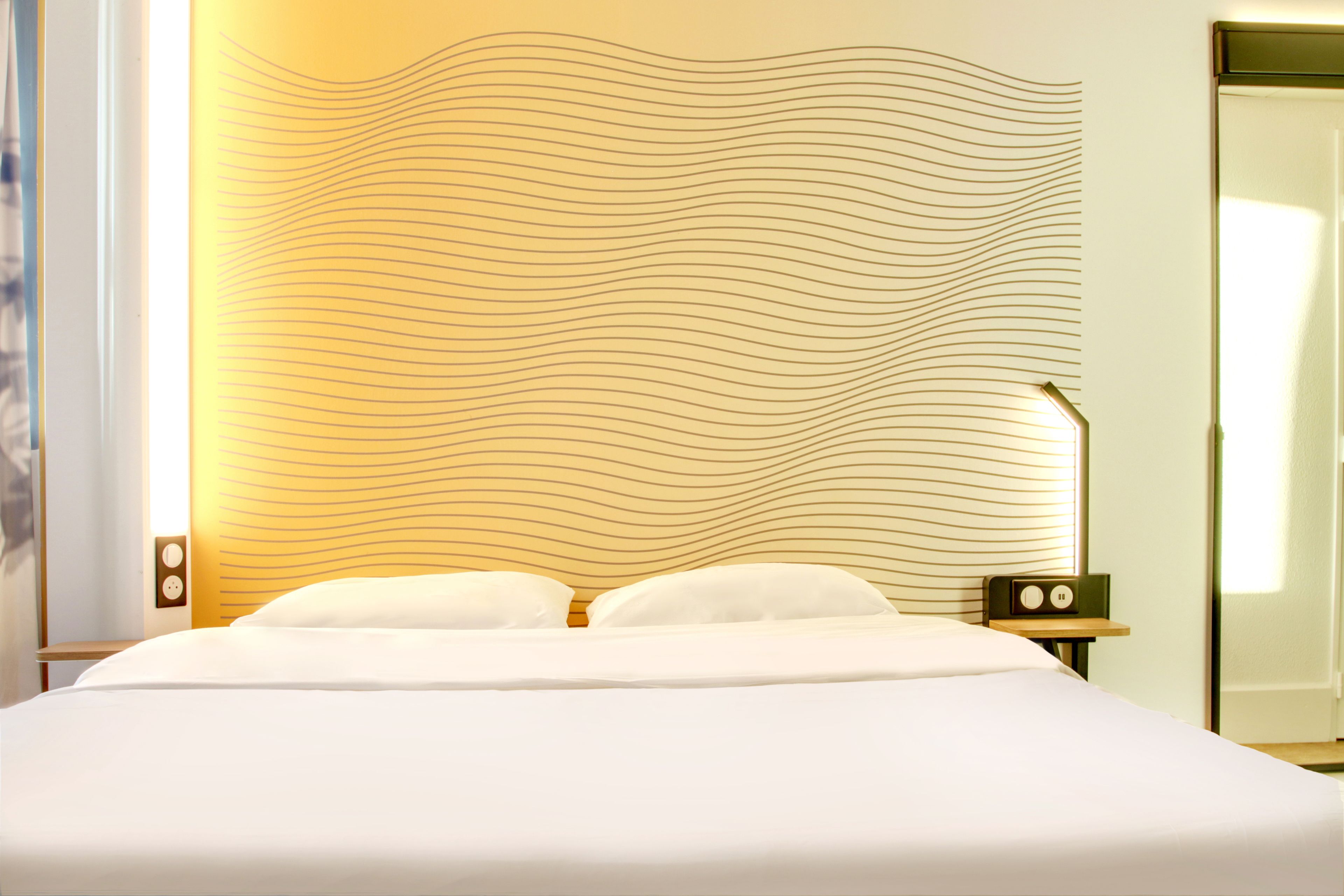 A neatly made bed with white linens, a wall with a wavy pattern, and a bedside table with a radio.