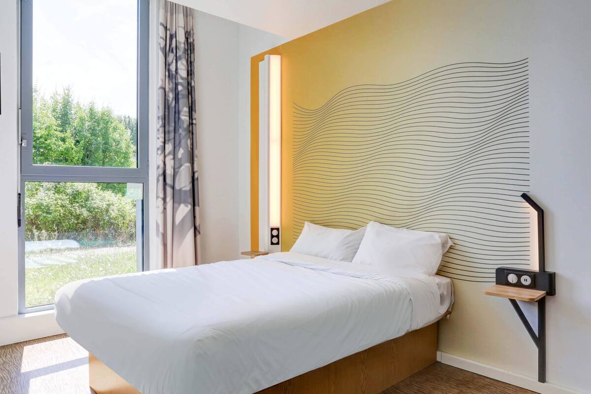 Quarto moderno com uma cama grande, mesa de cabeceira, luminária na parede e vista para o verde lá fora.