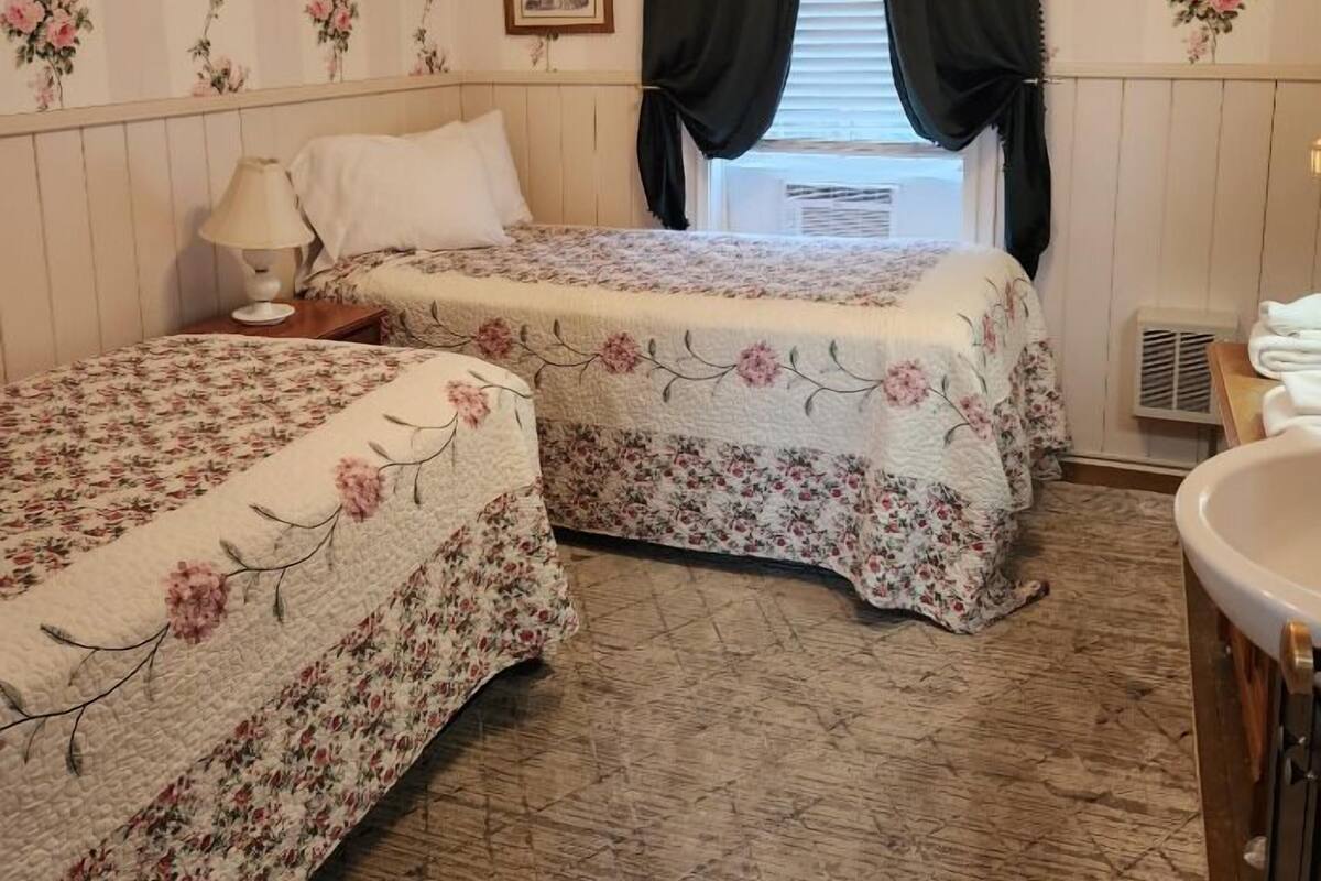 A bedroom with two beds, floral wallpaper, a picture on the wall, and a window with curtains.