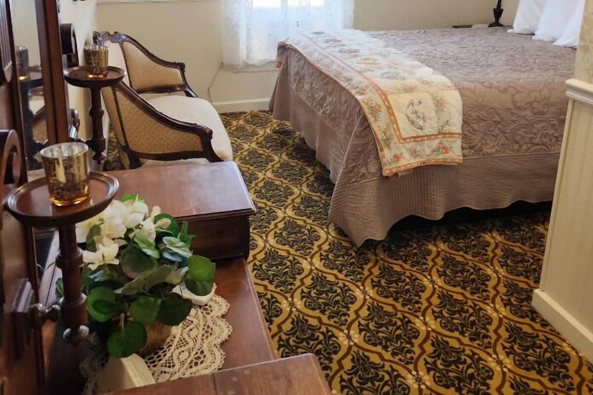 A bedroom with a bed, a chair, a table with a lamp, and a window with curtains.