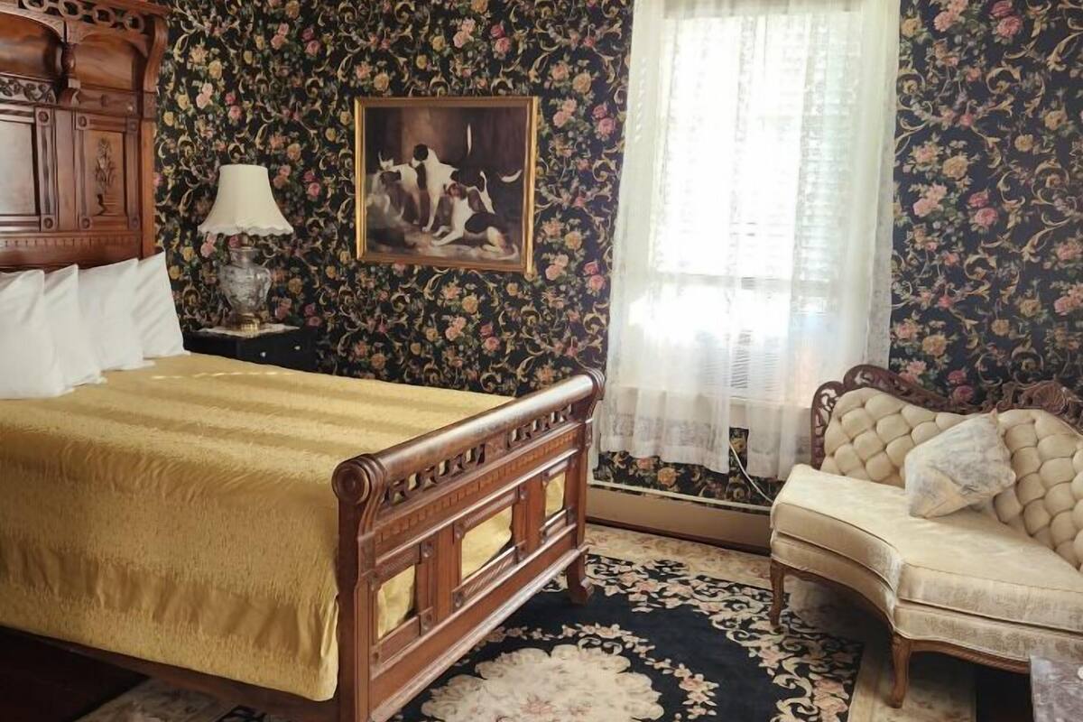 A bedroom with a wooden bed, a sofa, a floral-patterned wallpaper, a painting, and a window with sheer curtains.