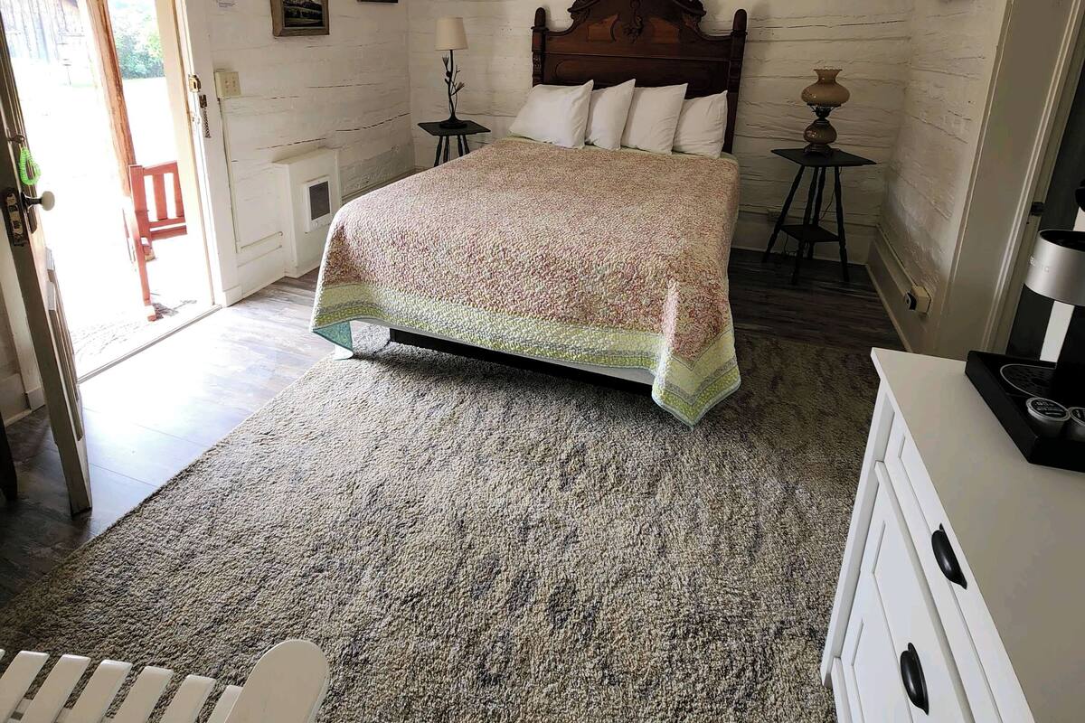 A bedroom with a wooden bed, a small bedside table with a lamp, a dresser, and a door leading to an outdoor area.