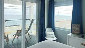A hotel room with a bed, a chair with a 'relax' pillow, a small table, and a view of the ocean through a large window. - The Grandview Hotel (York Beach)