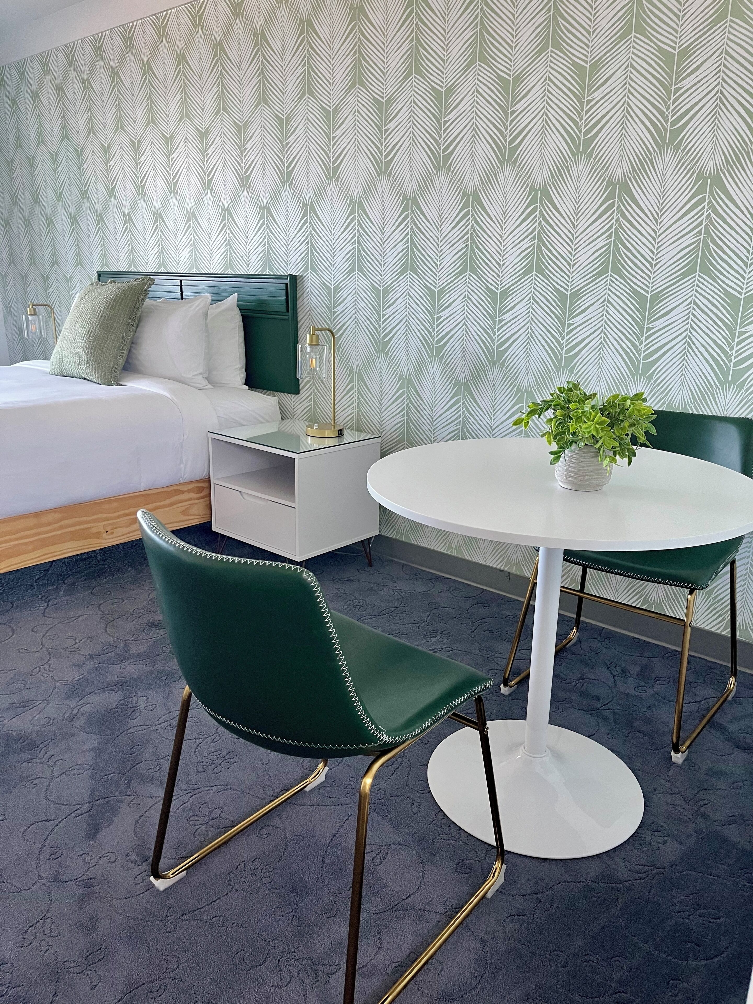 A modern hotel room with a bed, a bedside table with a lamp, a green upholstered chair, a round dining table with a white tablecloth, and a potted plant.