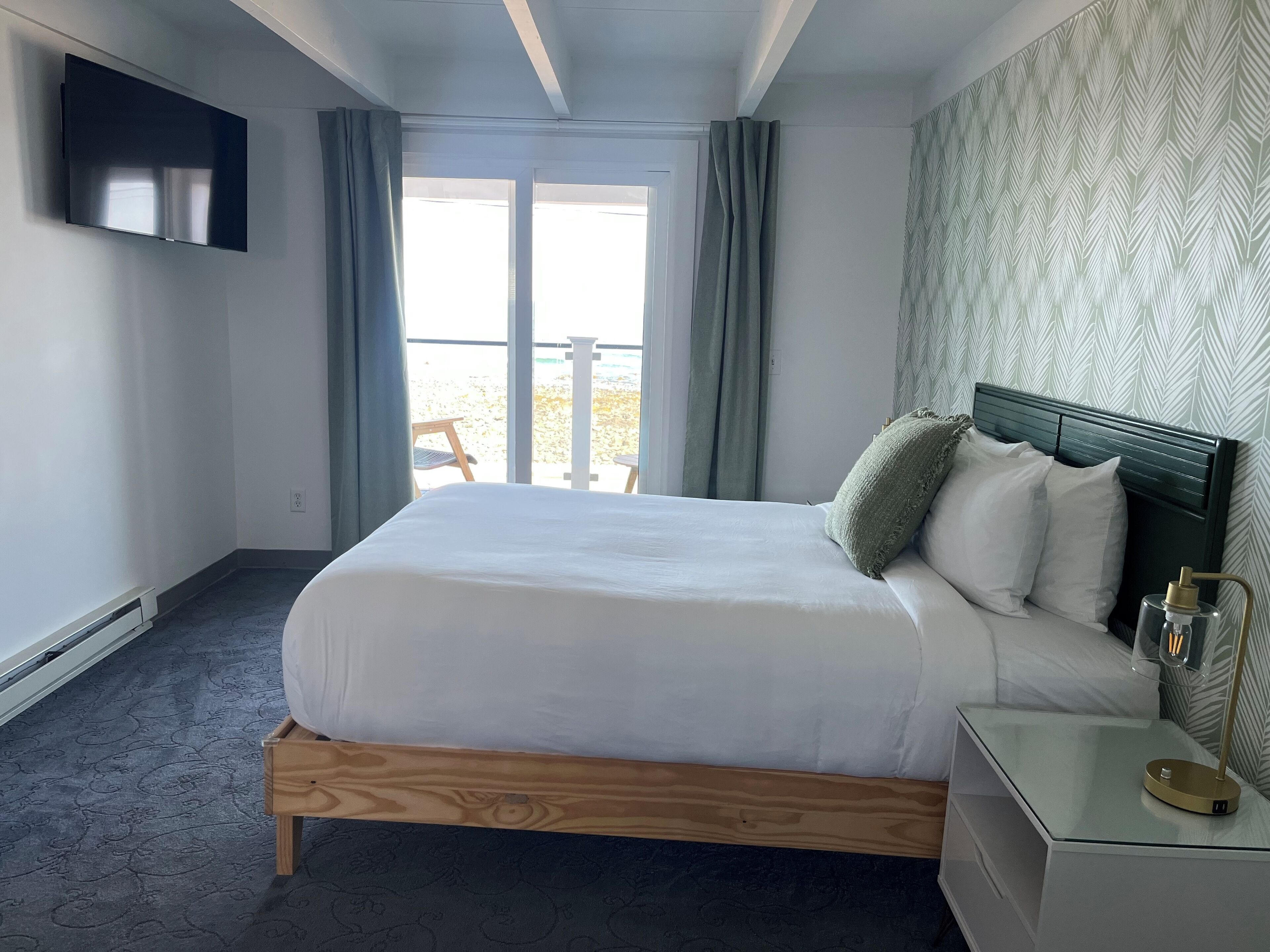 A hotel room with a bed, a bedside table with a lamp, a TV mounted on the wall, and a window with curtains.