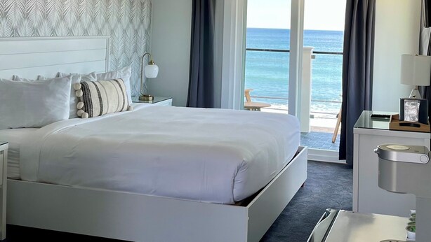 A hotel room with a large bed, a nightstand with a lamp, a desk with a lamp, a minibar, and a view of the ocean through a sliding door. - The Grandview Hotel (York Beach)