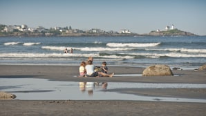 A beach with a family sitting on the sand, waves in the background, and a lighthouse in the distance. - The Grandview Hotel (York Beach)