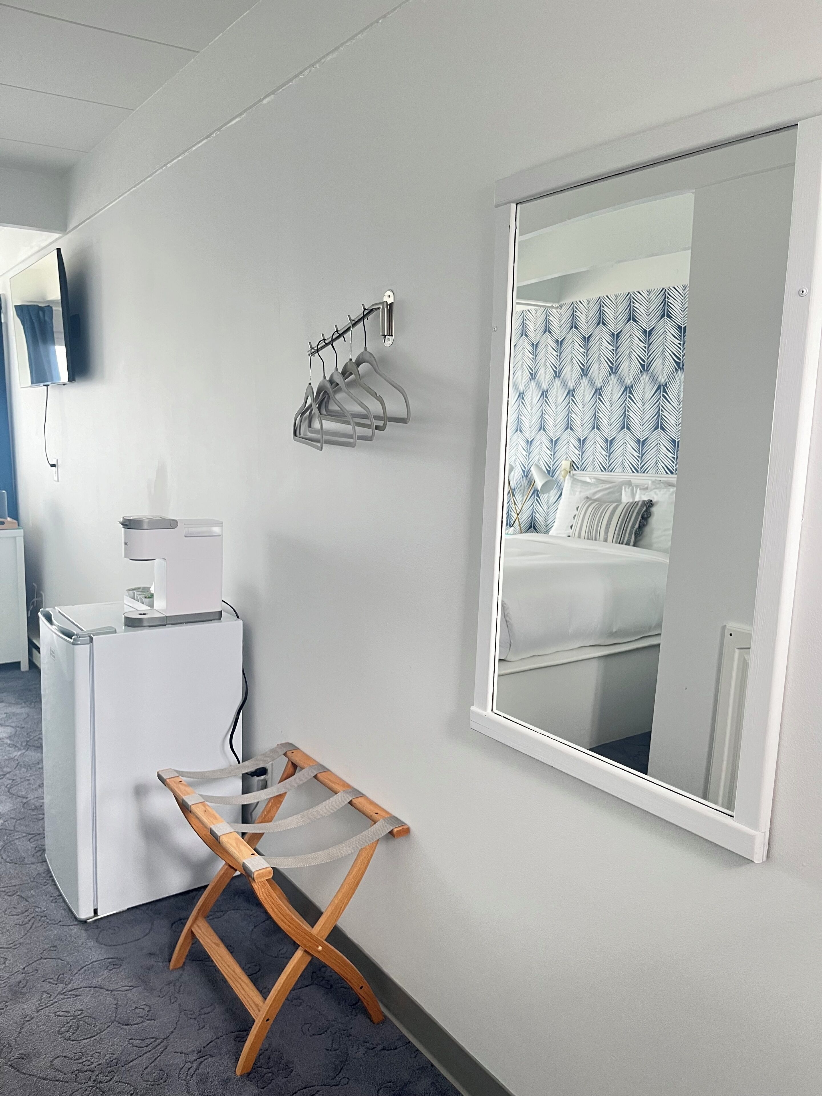 Quarto de hotel com cama, um espelho grande, varal fixado na parede, um eletrodoméstico pequeno e branco, e uma cadeira de madeira.